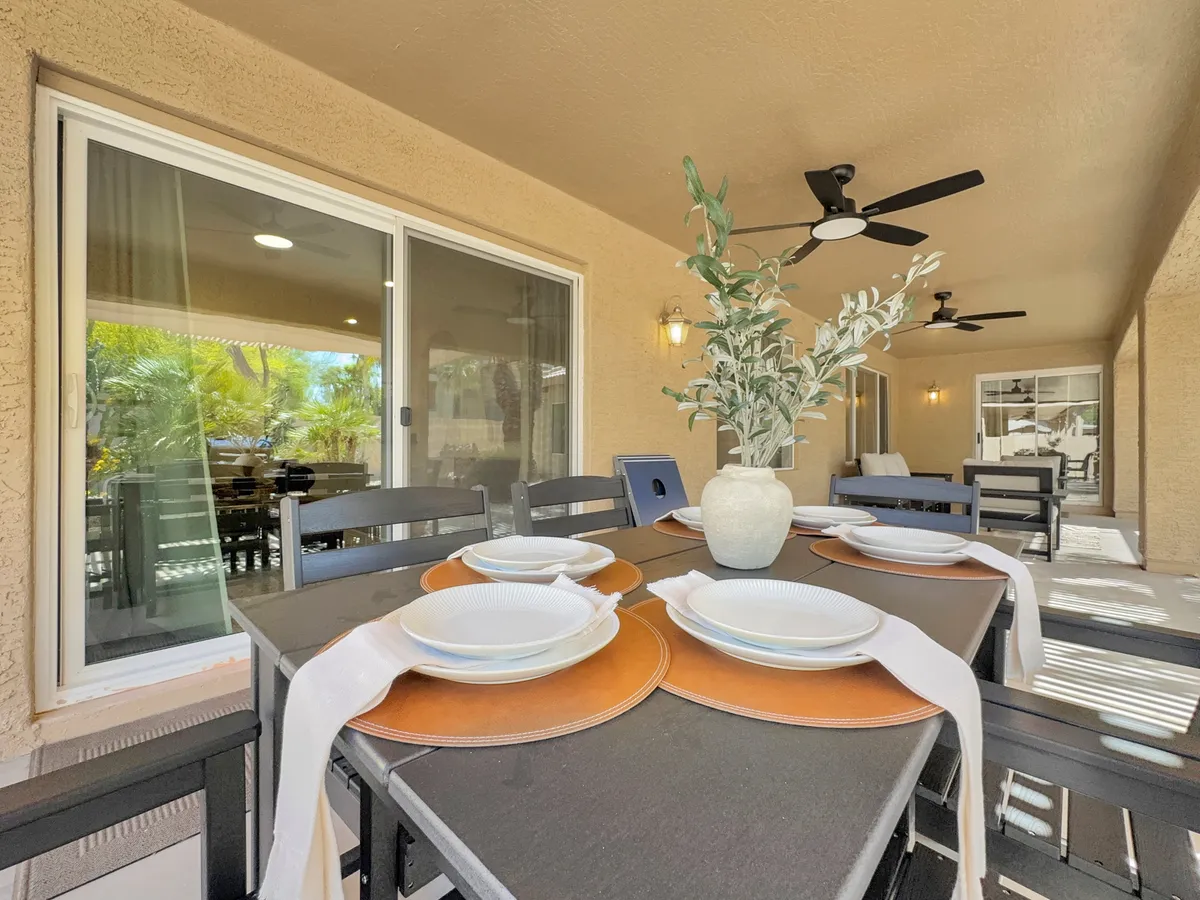 An inviting outdoor dining table set amid lush greenery, complete with plates, cutlery, and a decorative plant. Overhead outdoor ceiling fans suggest comfort even in sunny weather, and the scene evokes relaxation and social connection.