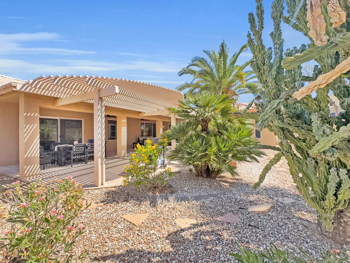 A tranquil backyard patio shaded by palm trees, with a covered seating area, desert landscaping, and a stone pathway winding through lush greenery. The scene captures an inviting spot for outdoor dining or relaxing in the Arizona sunshine, surrounded by peaceful garden views.