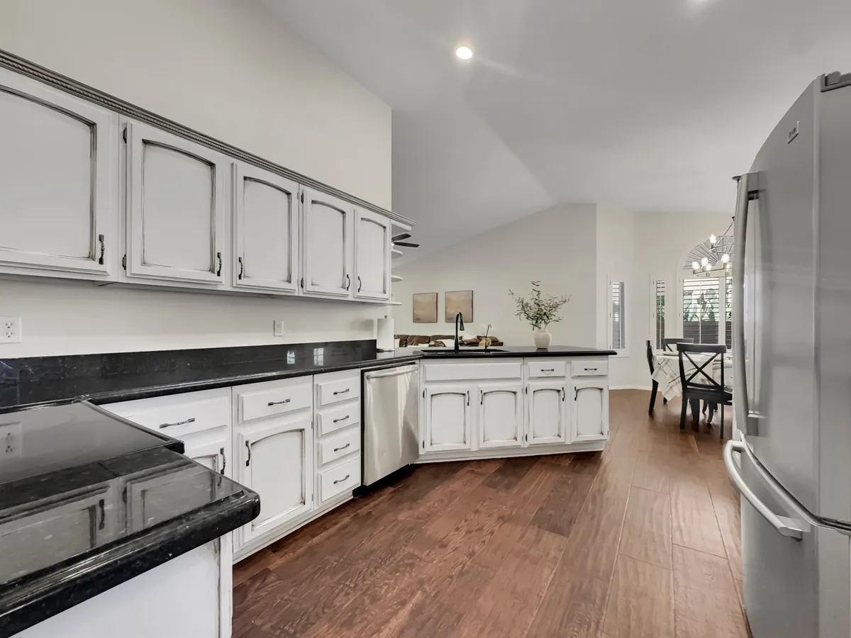 A spacious kitchen with black granite countertops and white cabinets, seamlessly flowing into a welcoming dining area in the background. The open-concept layout encourages social interaction while cooking and dining, with generous counter space and a welcoming mood ideal for family or group gatherings.
