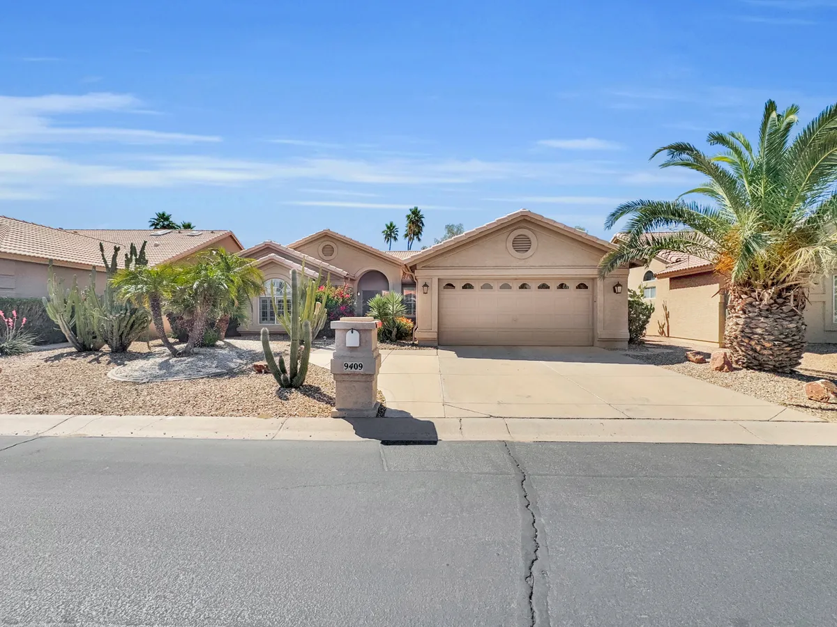 An exterior view of a home's front yard framed by palm trees and cacti, with a clear look at the house's facade. The landscaping hints at a relaxing, resort-like environment perfect for unwinding outdoors or enjoying a morning coffee on the porch.