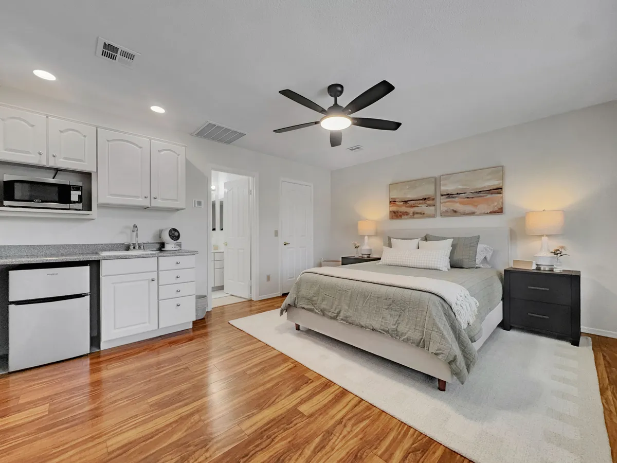 A comfortable bed adorned with decorative pillows forms the centerpiece, accompanied by a compact kitchenette area. Nightstands with lamps flank the bed, and inviting artwork hangs above. The layout suggests both comfort and autonomy, ideal for guests seeking extra privacy or an extended stay.