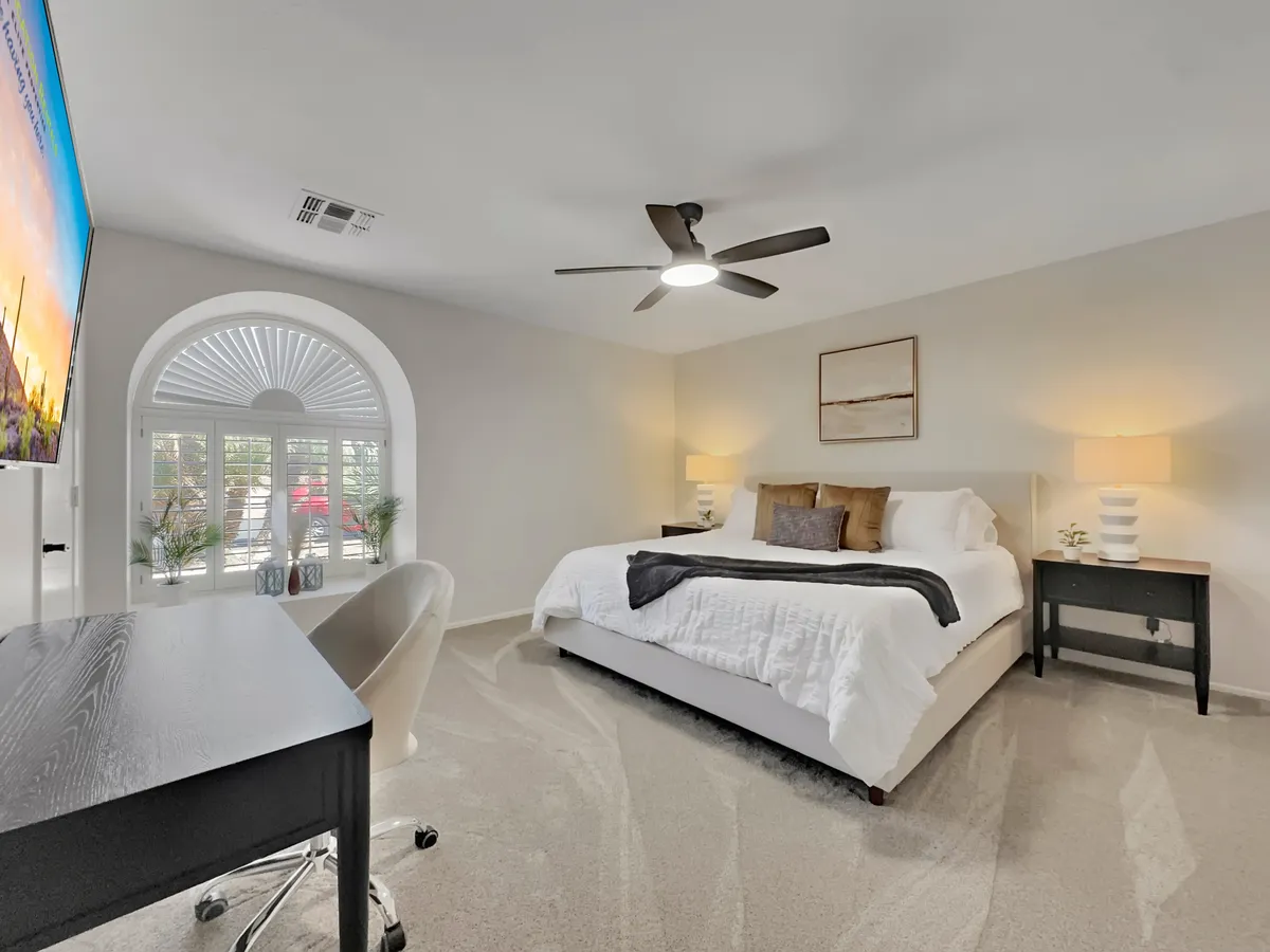 A spacious king-sized bed is positioned next to a large window, allowing plenty of natural light to filter in. The room includes a desk and chair, hinting at a functional workspace, while a ceiling fan and minimal decor create an airy, serene atmosphere. The space feels inviting and restful, perfect for relaxation or catching up on work.