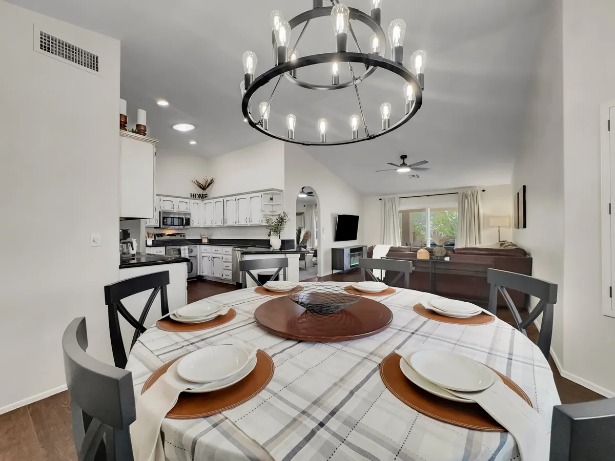 A bright open space seamlessly connecting the dining area, kitchen, and living room. The dining table is set and illuminated by a chandelier, with clear views of the adjacent living area, embodying the open, communal spirit.