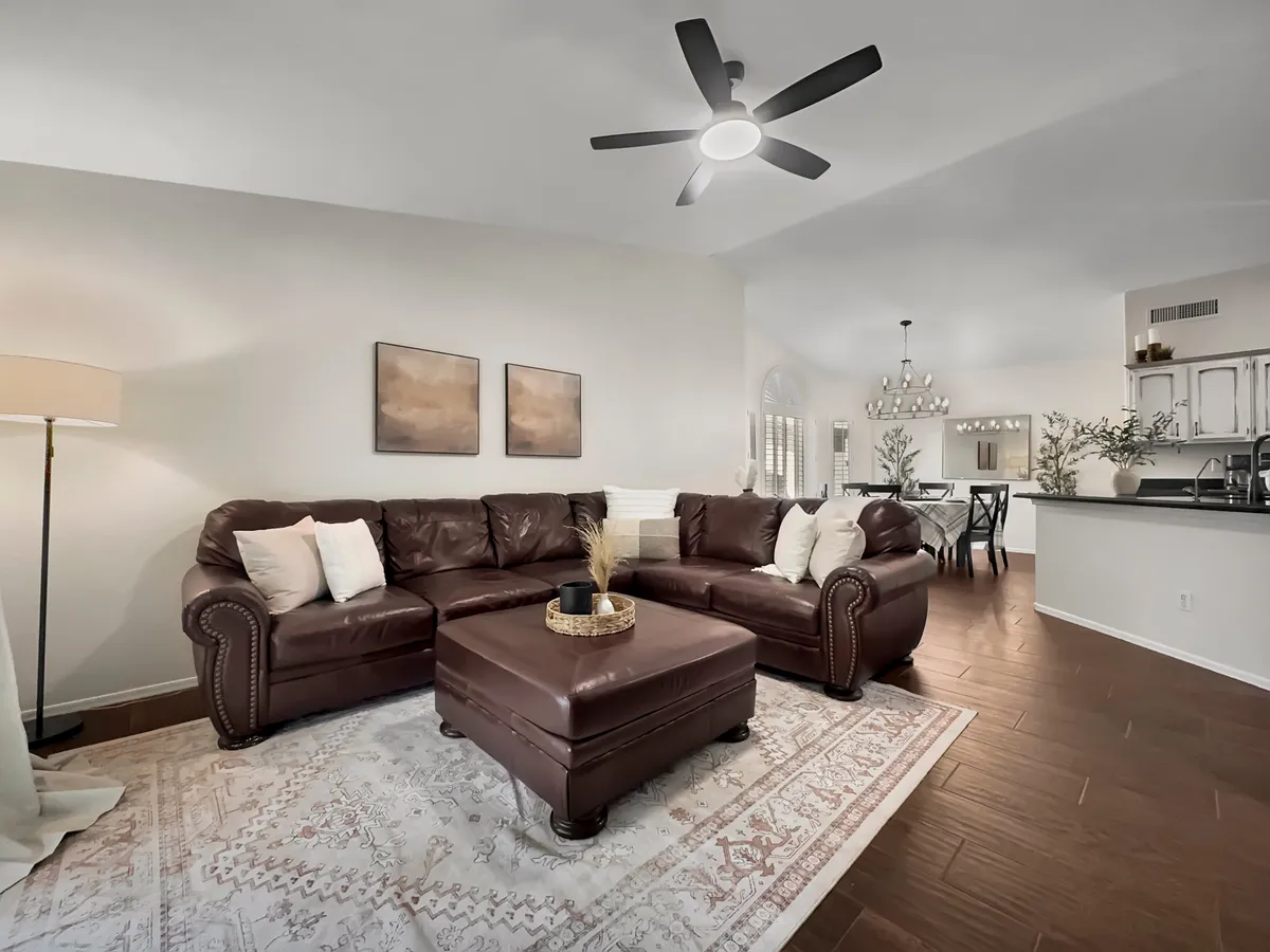 A spacious living room featuring a leather sectional sofa, coffee table, decorative rug, a floor lamp, and artwork on the wall. The room is bathed in natural light and arranged for both comfort and conversation, perfect for gatherings or relaxing evenings.