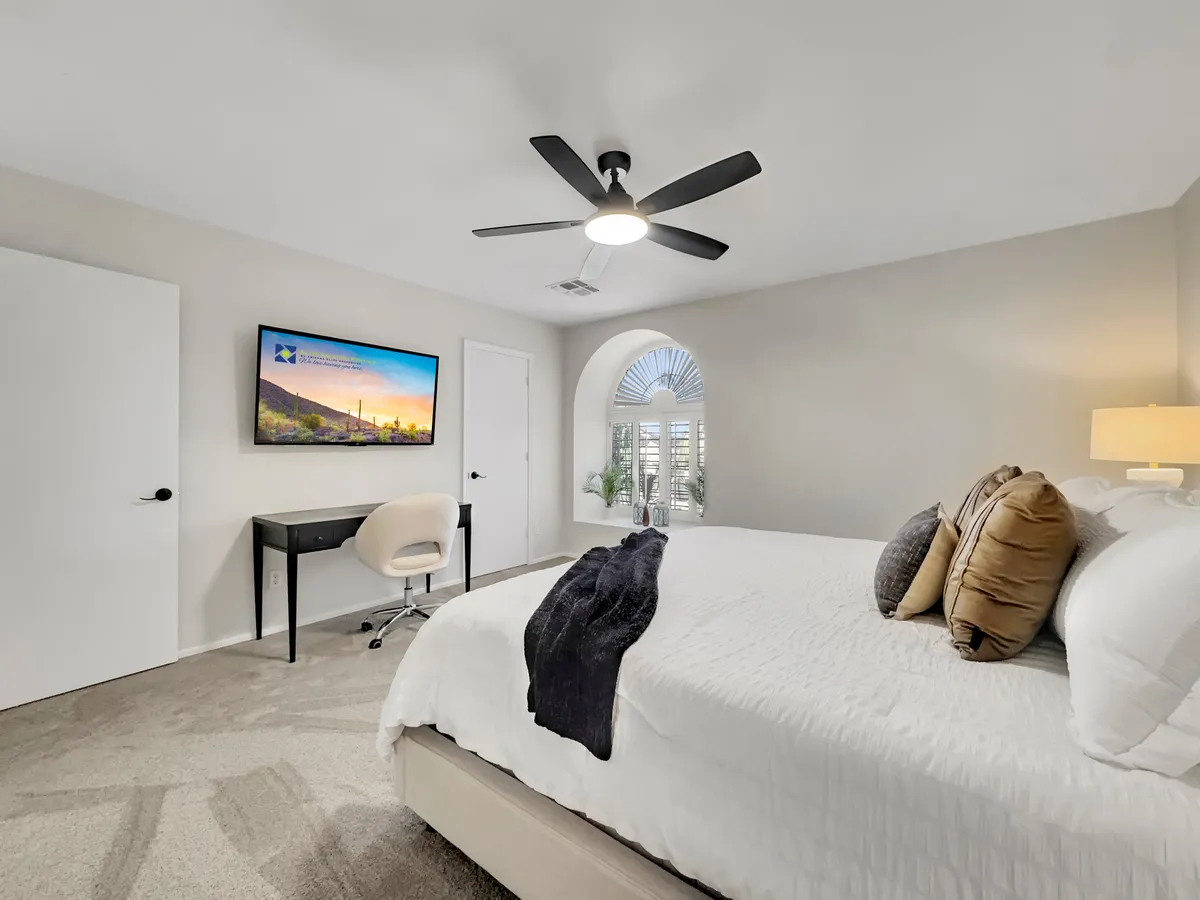 A cozy bedroom featuring a bed with decorative pillows, a TV mounted on the wall, and a desk and chair setup by an arched window with blinds. The space is filled with natural light, supporting both relaxation and productivity.