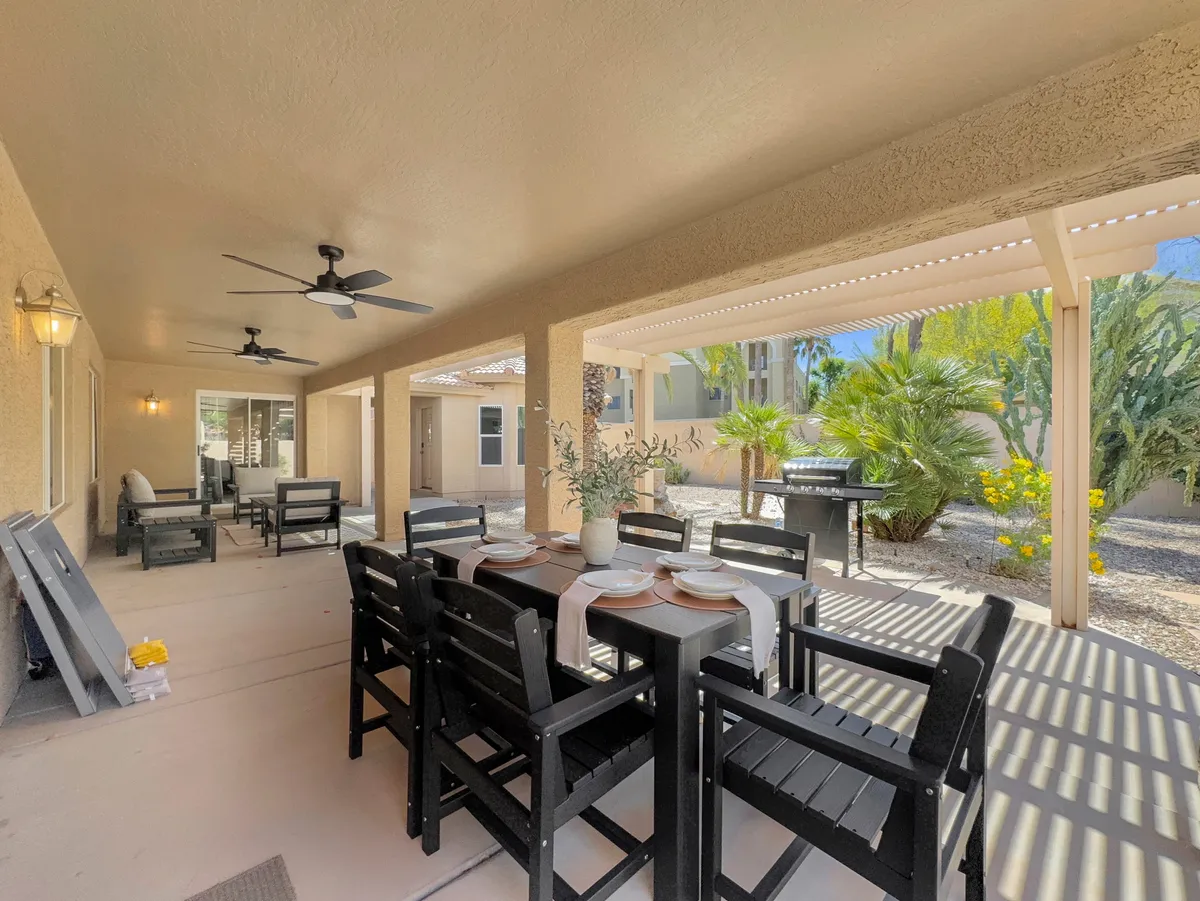 An exterior shot of a private patio area featuring a dining table set for a meal, comfortable seating, and lush landscaping with potted plants. The scene suggests a tranquil, resort-style space perfect for both relaxation and social gatherings.